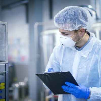 Technologist expert in protective uniform with hairnet and mask taking parameters from industrial machine in food production plant.